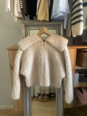 Krämvit teddyjacka med stor krage - Supermysig teddyjacka i krämvitt med oversized krage och dragkedja framtill. Jackan har en boxig passform och långa ärmar, perfekt för att hålla värmen. Tillverkad i fluffigt teddy-material som ger en trendig look.