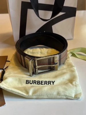 Burberry bälte - Snyggt bälte från Burberry med klassiskt randigt mönster i beige, brunt och rött. Bältet har en silverfärgad metallspänne och är tillverkat i skinn med textil på ovansidan. Perfekt accessoar för att lyfta din outfit.