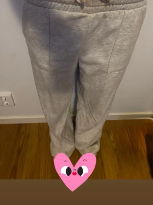 Ljusgrå mjukisbyxor med dragsko - Säljer ett par ljusgrå mjukisbyxor med raka ben och bred resår i midjan samt dragsko. Byxorna har två fickor framtill och är tillverkade i mjukt, bekvämt material som passar perfekt för chill dagar. Enkla att matcha med hoodie eller t-shirt.