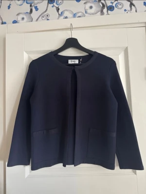 Mörkblå kofta från Isay - Mörkblå kofta från Isay med öppen front och rund hals. Koftan har två stora fickor framtill och är gjord i ett mjukt, stretchigt material. Enkel och clean design som funkar till det mesta.