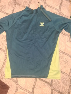 Blågrön träningströja från Hummel - Snygg långärmad träningströja från Hummel i blågrönt med ljusgula sidopaneler. Tröjan har halv dragkedja vid halsen och Hummel-logga på bröstet. Perfekt för träning eller uppvärmning. Tillverkad i ett lätt och stretchigt funktionsmaterial.