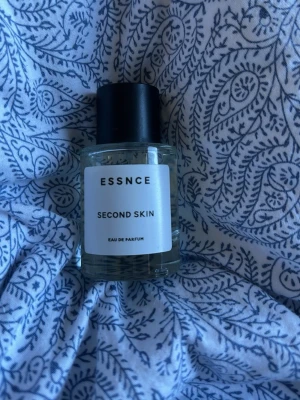 ESSNCE Second Skin Eau de Parfum - En stilren parfymflaska från ESSNCE med doften Second Skin. Flaskan är genomskinlig med en vit etikett och svart lock. Modern och minimalistisk design som passar perfekt på sminkbordet. Eau de Parfum-varianten ger en långvarig doft.