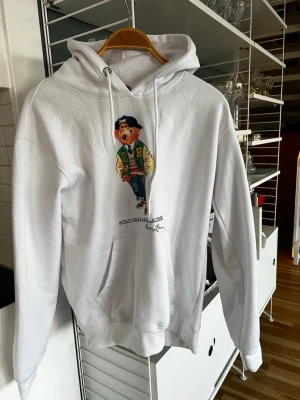Vit hoodie Polo Ralph Lauren björn - Säljer en vit hoodie från Polo Ralph Lauren med den ikoniska björnen på bröstet. Hoodien har huva med dragsko, magficka och ribbade muddar. Perfekt för dig som gillar streetwear och klassisk stil. Materialet är mjukt och skönt bomullstyg.