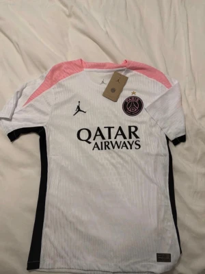 PSG matchtröja vit/rosa Jordan - Snygg PSG matchtröja i vitt med rosa axelpartier och svarta detaljer. Tröjan har Paris Saint-Germain-logga, Jordan-logga och Qatar Airways-tryck på bröstet. Perfekt för fotbollsträning eller att bära till vardags. Tillverkad i lätt och ventilerande material.
