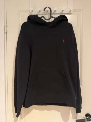 Svart hoodie från Polo Ralph Lauren - Storlek S, Klassisk svart hoodie från Polo Ralph Lauren med röd broderad logga på bröstet. Hoodien har stor huva, känguruficka framtill och ribbade muddar vid ärmslut och nederkant. Tillverkad i mjukt bomullsmaterial för en skön och avslappnad look.