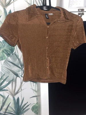 Brun kortärmad topp från H&M Divided - Säljer en brun kortärmad topp från H&M Divided i glansigt material. Toppen har krage, knappar framtill och en lätt croppad passform. Perfekt att styla med jeans eller kjol för en trendig look.