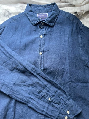 Marinblå linneskjorta Charles Tyrwhitt - Snygg marinblå skjorta från Charles Tyrwhitt i linne. Klassisk krage, vita knappar och lång ärm. Perfekt för dig som gillar en stilren och avslappnad look. Skjortan har en rak passform och känns lätt och luftig.