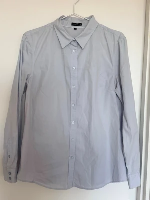 Ljusblå skjorta från Stockh LM - Klassisk ljusblå skjorta från Stockh LM med lång ärm och knappar framtill. Skjortan har en enkel, rak passform och är tillverkad i mjuk bomull. Perfekt till jeans eller kjol för en clean look. En liten reva på andra bilden, därav priset. 