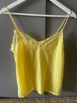 Gul spetsdetaljerad linnetopp - Snygg gul linnetopp med tunna axelband och spetsdetaljer längs urringningen. Perfekt för varma dagar och enkel att styla med jeans eller kjol. Luftigt material och feminin look.