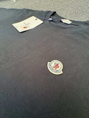 Svart Moncler t-shirt med logga - Snygg svart t-shirt från Moncler med rund halsringning och klassisk passform. På bröstet sitter Monclers ikoniska logotyp i vitt, blått och rött. Tillverkad i mjuk bomull som känns skön mot huden. Perfekt för dig som gillar stilrena och exklusiva plagg.