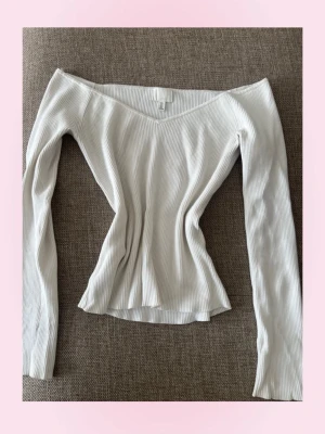 Vit ribbad offshoulder topp - Säljer en snygg vit ribbad topp med offshoulder-design och lång ärm. Toppen har en v-ringad hals och är figurnära med stretchigt material som ger en smickrande passform. Perfekt för dig som gillar stilrena och trendiga plagg.
