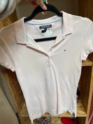 Ljusrosa pikétopp från Tommy Hilfiger - Klassisk ljusrosa pikétopp från Tommy Hilfiger med liten logga på bröstet. Toppen har krage, knappar framtill och korta ärmar. Tillverkad i mjuk bomull som känns skön mot huden. Perfekt för en clean och sportig look.