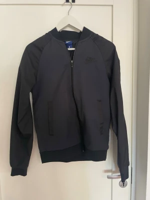 Svart bomberjacka från Nike - Snygg svart bomberjacka från Nike med diskret broderad logga på bröstet. Jackan har dragkedja framtill, två fickor och ribbade muddar vid ärmslut och nederkant. Perfekt för dig som gillar sportig och clean stil. 