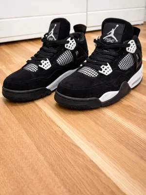 Svarta Air Jordan 4 Retro sneakers - Säljer ett par svarta Air Jordan 4 Retro sneakers med vita och grå detaljer. Skorna har en klassisk hög siluett, meshpaneler och ikoniska plastdetaljer vid snörningen. Ovandelen är i mocka och de har en bekväm sula med bra dämpning. Perfekta för dig som gillar streetwear.