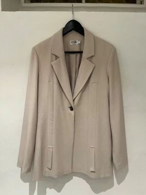 Beige kavaj från NA-KD  - Stilren beige kavaj från NA-KD med klassisk passform och ett knappstängning framtill. Kavajen har två fickor framtill och är tillverkad i ett mjukt, vävt material. Perfekt för en clean och modern look.