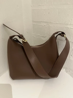 Brun axelväska i läderimitation - Snygg brun axelväska med bred axelrem och guldfärgade metalldetaljer. Väskan har en minimalistisk design och stängs med dragkedja. Perfekt för dig som gillar stilrena accessoarer och vill ha plats för det viktigaste.