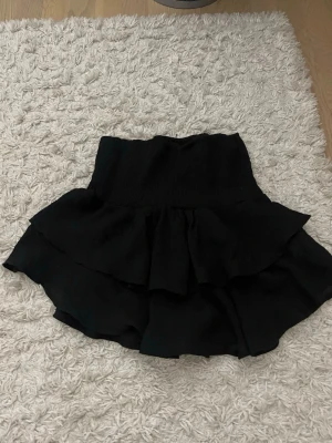 Svart volangskort - Supersnygg svart skort med volang. Kjoldesign framtill men shorts under, perfekt för en trendig och bekväm look. Tillverkad i ett mjukt, lätt tyg som ger skön känsla och snyggt fall.