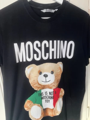 Svart Moschino t-shirt med björn - Säljer en svart t-shirt från Moschino med stort vitt logotryck och en söt björn på framsidan. Björnen har en tröja i grönt, vitt och rött med texten 'This is not a Moschino toy'. Klassisk passform och rund hals.