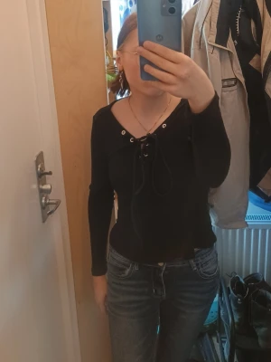 Svart långärmad topp med snörning - Snygg svart långärmad topp med snörning framtill och djupare ringning. Toppen har en slim passform och är perfekt att styla med jeans eller kjol. Materialet känns mjukt och stretchigt, troligen i bomull eller polyesterblandning.