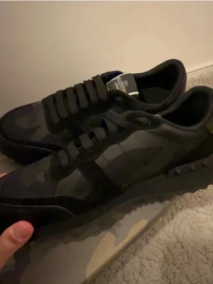 Svarta rockrunners från Valentino - Snygga svarta rockrunners från Valentino med kamouflagemönster och detaljer i mocka och textil. Skorna har svarta skosnören och en låg siluett som ger en clean och modern look. Perfekta för dig som gillar exklusiva streetwear-vibbar.