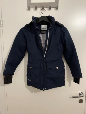Mörkblå parkas från McKinley - Mörkblå parkas från McKinley med avtagbar huva, dragkedja och tryckknappar framtill. Jackan har stora fickor med lock, muddar vid ärmslut och är fodrad för extra värme. Perfekt för kalla dagar och outdoor-äventyr.