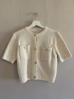 Kortärmad vit kofta med fickor - Stilren vit kofta med korta ärmar och rund hals. Koftan har två bröstfickor med dekorativa knappar och stängs framtill med fyra pärlemorskimrande knappar. Materialet är stickat med diskret struktur för en trendig look.