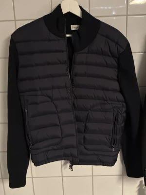 Svart quiltad cardiganjacka Moncler - Svart cardiganjacka från Moncler med quiltad front och släta ärmar. Jackan har dragkedja framtill, två fickor med dragkedja och en broderad Moncler-logga på ärmen. Materialmix med vadderad kropp och stretchiga ärmar för en clean och modern look.