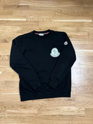 Svart sweatshirt från Moncler - Säljer en svart sweatshirt från Moncler med stort vitt logomärke på bröstet och en mindre patch på ärmen. Tröjan har rund halsringning och ribbade muddar vid ärmslut och nederkant. Perfekt för dig som gillar streetwear och exklusiva märken.