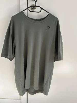 Oversized grön t-shirt från Gymshark - Snygg oversized t-shirt i militärgrönt från Gymshark. T-shirten har rund hals, korta ärmar och stor svart logga på ryggen samt liten logga framtill. Tillverkad i mjukt bomullsmaterial, perfekt för gymmet eller chill. 