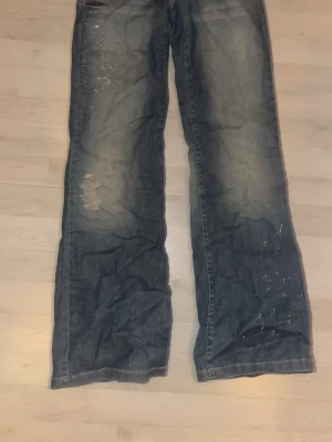 Blå bootcut jeans med slitningar - Säljer ett par blå bootcut jeans med slitningar och ljusa partier framtill. Jeansen har dekorativa sömmar på bakfickorna och en brun läderpatch i midjan. Materialet är klassisk denim och passformen är avslappnad med utsvängda ben.