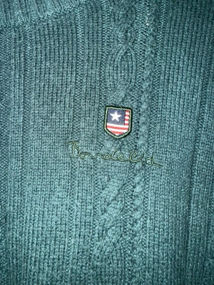 Bondelid stickad tröja  - Mörkgrön stickad tröja  med klassiskt mönster och liten broderad logga på bröstet. Rund hals och skön passform. nopprig.