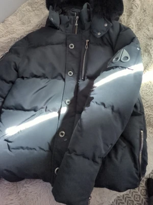 Svart dunjacka från Moose Knuckles - Säljer en svart dunjacka från Moose Knuckles med avtagbar huva och fuskpäls. Jackan har flera dragkedjeförsedda fickor, stora knappar och logotyp på ärmen. Perfekt för kalla vinterdagar tack vare tjockt dunfoder och vindtätt material.