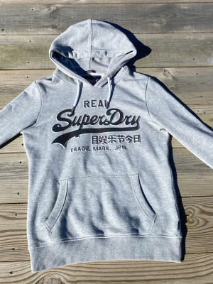 Grå hoodie från Superdry - Snygg grå hoodie från Superdry med svart logga och japansk text på bröstet. Perfekt för en avslappnad look. Tröjan är limited edition, alltså går den inte att köpa i butik. Tröjan är ytterst lite använd, endast ett fåtal gånger. Pris går att diskuteras!!
