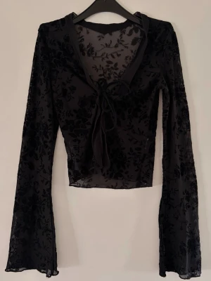 Svart mesh blus med knyt och blommönster - Snygg svart mesh blus med transparent tyg och sammetsliknande blommönster. Blusen har långa utsvängda ärmar och knyt framtill som ger en trendig look. Perfekt att styla med höga jeans eller kjol.