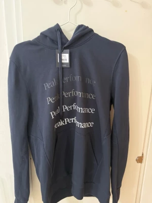 Mörkblå hoodie från Peak Performance - Snygg mörkblå hoodie från Peak Performance med tryckt logga i flera storlekar på bröstet. Klassisk huva med dragsko och stor magficka framtill. Tillverkad i mjukt material som känns skönt mot huden. Perfekt för en avslappnad och sportig stil.
