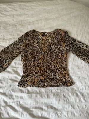 Mönstrad blus med knappar - Snygg blus med djurmönster i beige, brunt och svart. Djup v-ringning och dekorativa knappar framtill. Figurnära passform och långa ärmar. Perfekt för dig som vill sticka ut med ett trendigt mönster. Materialet känns lätt och luftigt.