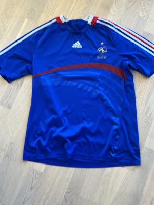 Blå Frankrike Adidas fotbollströja VM 2006 - Säljer en blå fotbollströja från Adidas med Frankrikes landslagsmärke och en stjärna broderad på bröstet. Tröjan har vita och röda detaljer på axlarna samt en röd diagonal linje över framsidan. Tillverkad i lätt och ventilerande material.