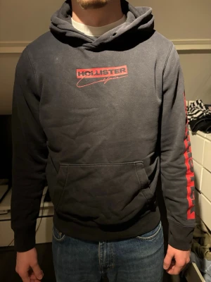 Svart Hollister hoodie med röd logga - Snygg svart hoodie från Hollister med röd logga på bröstet och stort HOLLISTER-tryck längs ena ärmen. Klassisk känguruficka framtill och huva. Perfekt för dig som gillar streetwear och vill ha något bekvämt och stilrent. Materialet känns mjukt och skönt.