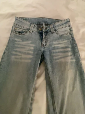 Ljusblå jeans från Cheep monday - Säljer ett par ljusblå jeans med slitningar framtill och en unik grön patch med tryck bak i midjan. Klassisk femficksmodell och raka ben. Jeansen har en cool streetwear-vibe och är tillverkade i denim.