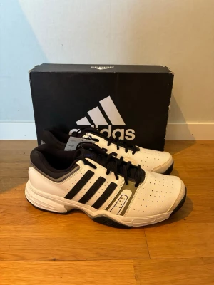 Adidas tennis skor  - Passar även storlek 44                         Snygga vita sneakers från Adidas med klassiska svarta ränder på sidorna och svart sula. Skorna har perforerade detaljer för ventilation, svart innersula och snörning. Perfekta för sport eller streetwear. Materialet är en mix av syntet och textil.