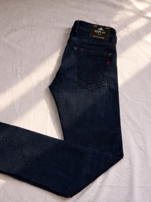 Mörkblåa replay grover jeans - Snygga mörkblå jeans från Replay i slim fit-modell. Jeansen har klassisk femficksdesign, subtila slitningar och en liten röd detalj på bakfickan. Tillverkade i mjukt denimtyg som ger en skön känsla och bra. Kom pm vid fundering.  A: 42 B 81 C 23 D 20 E 19