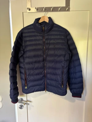 Mörkblå pufferjacka med dragkedja - Snygg ralph lauren jacka. Jackan är inte speciellt tjock. Perfekt för vår, höst och sommarkvällar.  Jackan har en söm som har släppt cirka 4 cm stort hål på undersidan. Påverkar inte dunet i jacken eller fickan. Vänstra fickans dragkedja är även sönder där av priset. (Nypris runt 2600) Pris går att diskutera, hör av er vid minsta funderingen😊