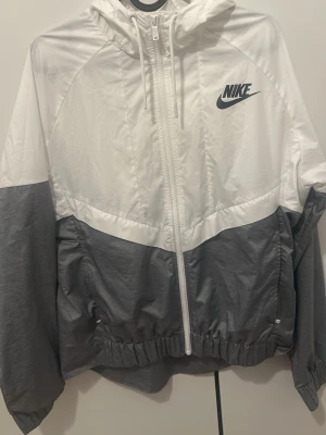 Nike vit och grå vindjacka - Nike vindjacka i vitt och grått med dragkedja framtill, huva och elastiska muddar. Jackan har en sportig look med logga på bröstet och två sidofickor. Tillverkad i lätt, vindtätt material som passar perfekt för blåsiga dagar.