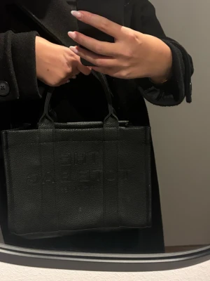 Svart The Tote Bag från Marc Jacobs ej äkta - Säljer en svart The Tote Bag från Marc Jacobs i präglat läder. Väskan har två korta handtag och en boxig, strukturerad form med diskret logga framtill. Perfekt storlek för det viktigaste och ger en clean, stilren look.