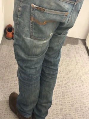 Blå jeans från Nudie Jeans med sjuk tvätt - Säljer ett par klassiska blå jeans från Nudie Jeans med slitningar och detaljerade sömmar på bakfickorna. Jeansen har en rak passform och är tillverkade i denim med en snygg tvättad look. Perfekta för en avslappnad och trendig stil. Mått: midja 42cm, längd 98cm, benöppning 19cm. Hör av er vid frågor och funderingar! Pris går att diskutera