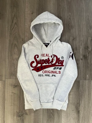 Ljusgrå hoodie från Superdry - Ljusgrå hoodie från Superdry med stor röd och svart logga på bröstet och tryck på ärmen🤩 passar storlek Xs och inga fel😇 Hör av er vid funderingar samt snabbaffär🙌🏻