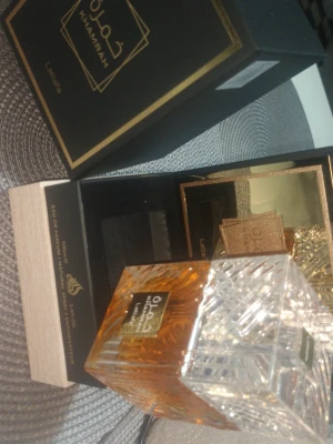 Lattafa Khamrah Eau de Parfum - Lattafa Khamrah Eau de Parfum kommer i en elegant fyrkantig glasflaska med ett detaljerat, kristalliknande lock. Flaskan har en varm bärnstensfärgad vätska och etikett i guld. Förpackningen är svart med guldiga detaljer och arabiska inskriptioner.