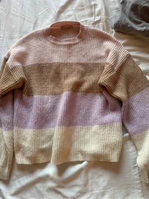 Randig stickad tröja från ONLY - Mysig stickad tröja från ONLY med breda ränder i beige, ljuslila, brun och ljusrosa. Tröjan har rund halsringning och långa ärmar. Perfekt för dig som gillar mjuka pasteller och en avslappnad stil.