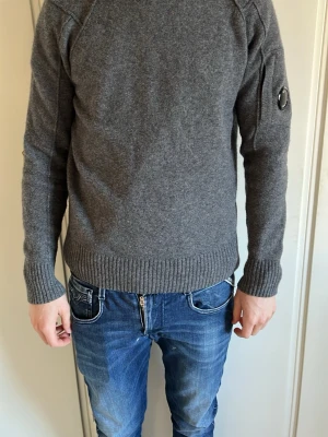 Grå stickad tröja från C.P. Company - Snygg grå stickad tröja från C.P. Company med rund hals och ribbade muddar. Tröjan har en ikonisk linsdetalj på ena ärmen och är tillverkad i mjukt material som passar perfekt till jeans. Modellen är 180 cm lång och väger 63 kg. Det är bara att höra av sig vid frågor och funderingar 😀