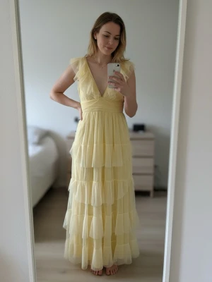 Ljusgul långklänning med volanger - Romantisk ljusgul långklänning med djup v-ringning, volangdetaljer och flera lager i tyll. Klänningen har ärmlös design och markerad midja för en feminin siluett. Perfekt för sommarens alla tillfällen.
